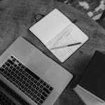 Black and white top view of a workspace with a laptop, notebooks, and pens on a wooden table.