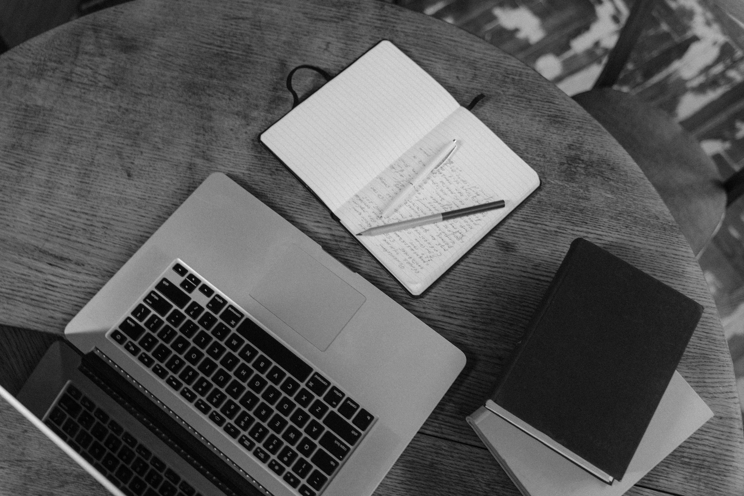 Black and white top view of a workspace with a laptop, notebooks, and pens on a wooden table.